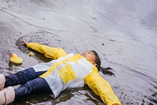 Високий кут наповненого азіатського хлопчика в гумових чоботах і ковток лежить у бридкій калюжі в дощовий день. — стокове фото