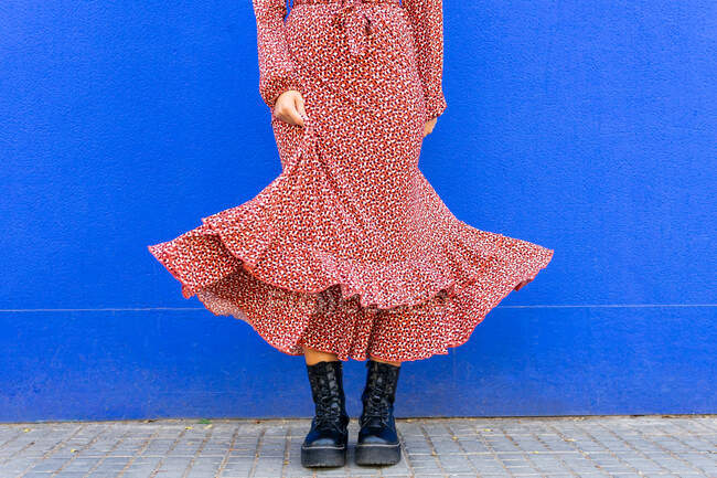 Нерозбірлива жінка в стильній червоній сукні стоїть на тротуарі до синьої стіни вдень. — стокове фото