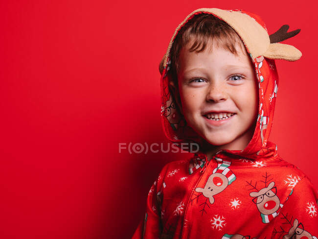 Очаровательный маленький мальчик в рождественской пижаме в капюшоне с оленем, стоящим на красном фоне и смотрящим в камеру — стоковое фото
