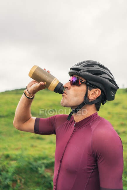 Vista lateral do esportista no capacete de ciclismo e óculos de sol bebendo água da garrafa durante a pausa do treino — Fotografia de Stock