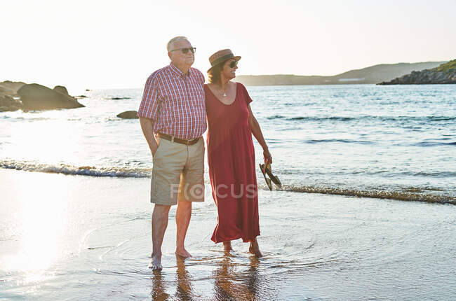 Cuerpo completo de sonriente pareja de ancianos descalzos en gafas de sol de pie en la playa de arena húmeda y disfrutando de un día soleado - foto de stock