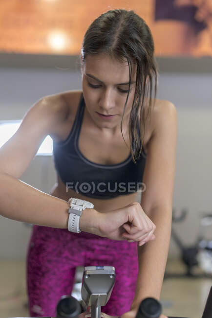 Сконцентрированная потная молодая спортсменка в топе и леггинсы, занимающаяся кардиотренировкой на велотренажере в тренажерном зале, проверка умных часов — стоковое фото
