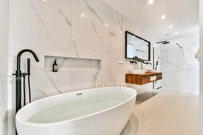 Pure water in oval shaped soaking tub against washstand under mirror and shiny lamps in contemporary bathroom in house — Stock Photo