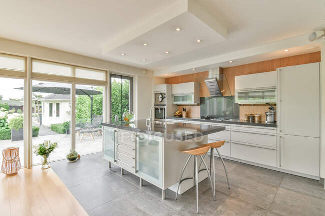 Moderne Kücheneinrichtung mit Tisch und Hockern gegen Kühlschrank und Schränke im Haus mit Glaswand tagsüber — Stockfoto
