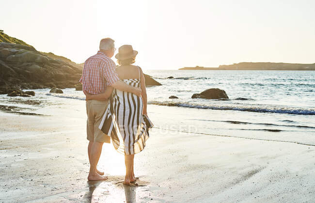 Vista trasera de pareja de ancianos descalzos irreconocibles en gafas de sol de pie en la playa de arena húmeda y disfrutando de un día soleado - foto de stock