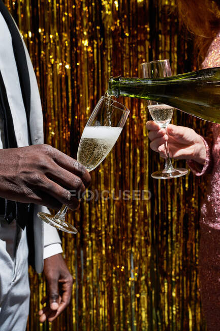 Crop mulher anônima derramando champanhe de garrafa em vidro de namorado preto durante a celebração da véspera de Ano Novo contra ouropel — Fotografia de Stock