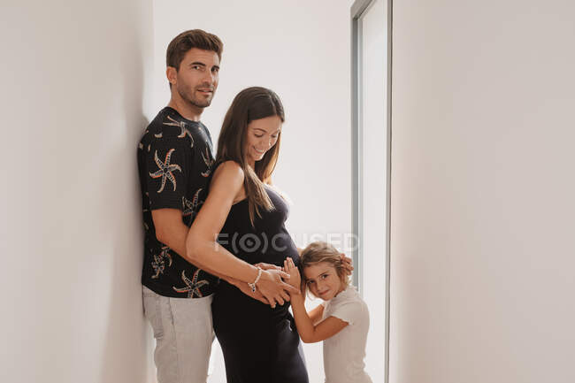 Vista lateral del niño acariciando el vientre de la sonriente futura madre contra papá mientras está de paso en casa - foto de stock