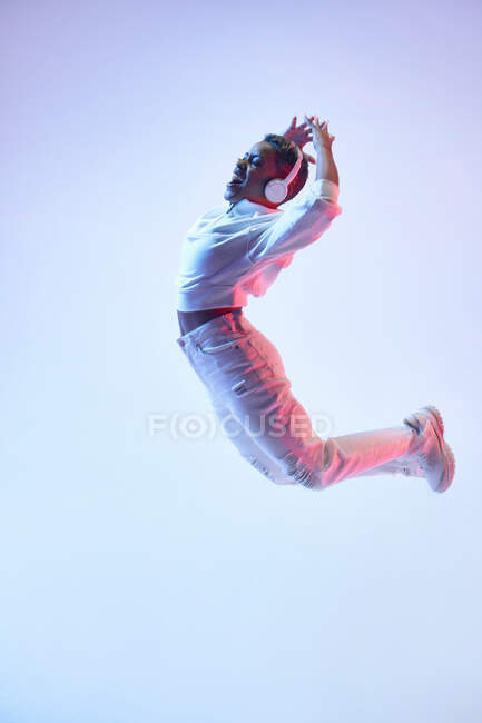 Vista lateral de mujer étnica enérgica en auriculares inalámbricos y ropa de moda saltando con la boca abierta mientras baila hip hop - foto de stock