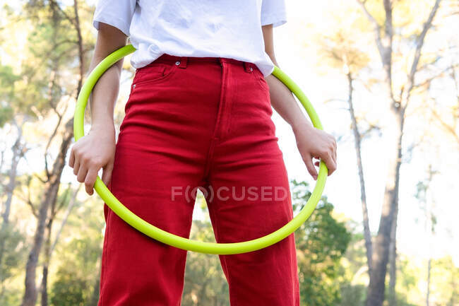 Desde abajo de la cosecha irreconocible adolescente en pantalones vaqueros rojos girando hula hoop mientras tiene tiempo libre en el parque - foto de stock