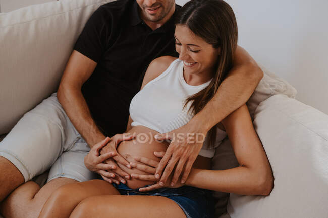 Розпещений чоловік обнімає животик вагітної коханої жінки, відпочиваючи на дивані у вітальні. — стокове фото