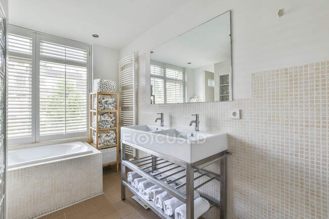 Белая ванна, установленная у окна в стильной ванной комнате с чистыми раковинами под зеркалом и бежевыми плиточными стенами в современной квартире днем — стоковое фото