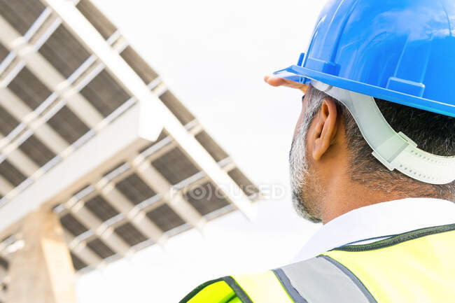 Vista trasera de un contratista masculino étnico maduro irreconocible recortado en chaleco y sombrero blanco parado cerca de la estación de energía solar - foto de stock