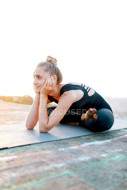 Corps complet de femme en forme en vêtements de sport pratiquant Ardha Padmasana et touchant le visage pendant l'entraînement de yoga en plein air au coucher du soleil — Photo de stock
