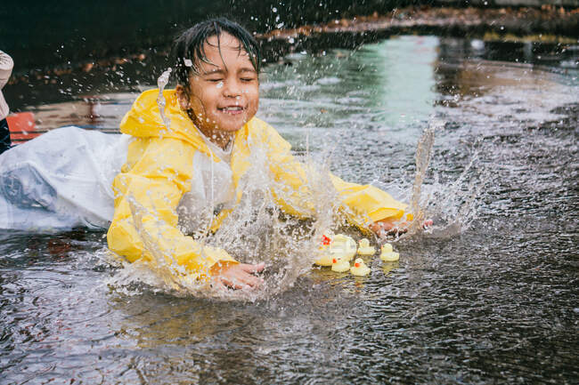 Encantador niño étnico en impermeable jugando con patos de plástico que se reflejan en charco ondulado en tiempo lluvioso - foto de stock