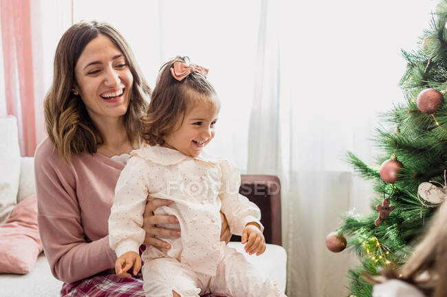 Mãe alegre abraçando a menina encantadora enquanto olha para longe contra o abeto decorado durante o feriado de Ano Novo no quarto da casa — Fotografia de Stock