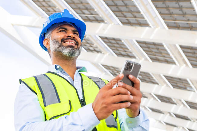 From below happy middle aged Hispanic foreman in in hardhat and waistcoat browsing on smartphone looking at screen while standing near solar power station — Stock Photo