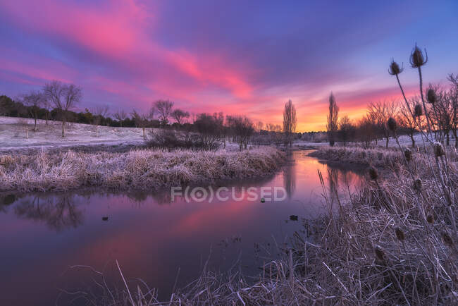 Scenery view of calm river flowing through grassy meadow covered with frost under bright sunset sky — Stock Photo