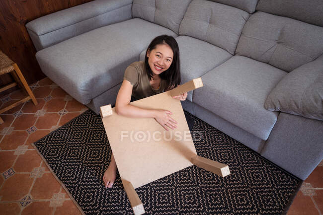 Молодая внимательная этническая женщина монтажный стол на декоративном ковре против дивана в светлом доме комнате — стоковое фото
