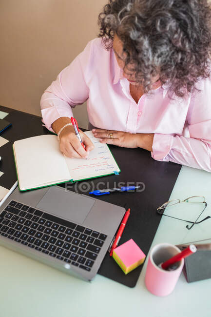 Alto ángulo de cultivo mujer emprendedora tomando notas en la agenda mientras trabaja en el escritorio con netbook en el espacio de trabajo - foto de stock
