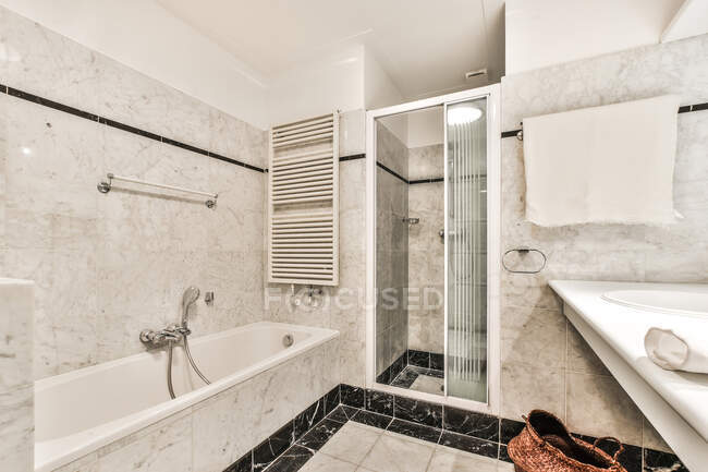 Интерьер современной белой кафельной ванной комнаты с ванной и душевой кабиной возле раковины под зеркалом и полотенцем — стоковое фото