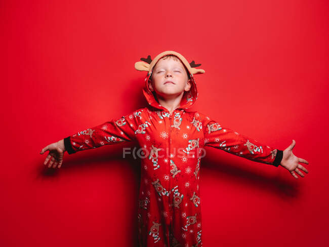 Adorable niño en pijama de Navidad con capucha con ciervos de pie sobre fondo rojo con los ojos cerrados y los brazos extendidos - foto de stock