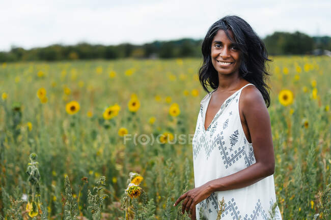 Aufrichtige erwachsene ethnische Frau blickt in die Kamera auf einer Wiese mit blühenden Blumen in der Landschaft auf verschwommenem Hintergrund — Stockfoto