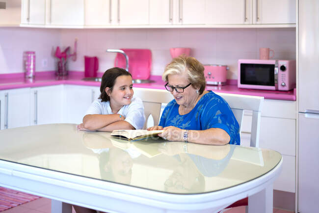 Grand-mère concentrée en vêtements décontractés et lunettes assis à table et livre de lecture avec petite-fille joyeuse dans la cuisine à la maison — Photo de stock