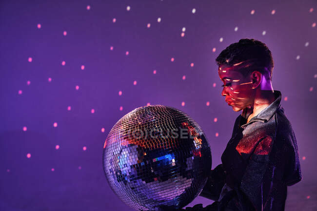 Vista laterale della giovane donna afroamericana alla moda con trucco e palla glitter in luce viola — Foto stock
