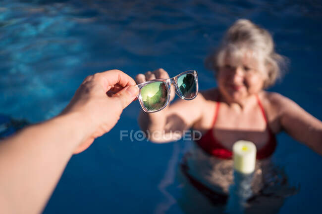 Сверху кукурузник дарит солнцезащитные очки пожилой женщине в бикини, плавающей в бассейне — стоковое фото