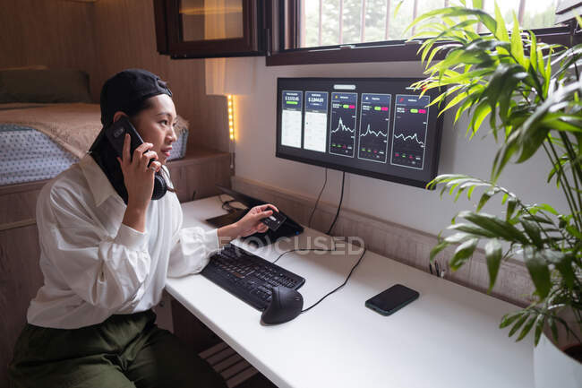 Surpreendida Asiática comerciante feminino falando no smartphone e olhando para o cartão de crédito enquanto trabalhava com criptomoeda estatística — Fotografia de Stock