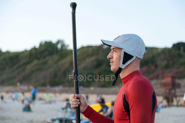 Seitenansicht eines männlichen Surfers in Neoprenanzug und Hut, der mit Paddel und SUP-Brett wegschaut, während er sich auf das Surfen an der Küste vorbereitet — Stockfoto