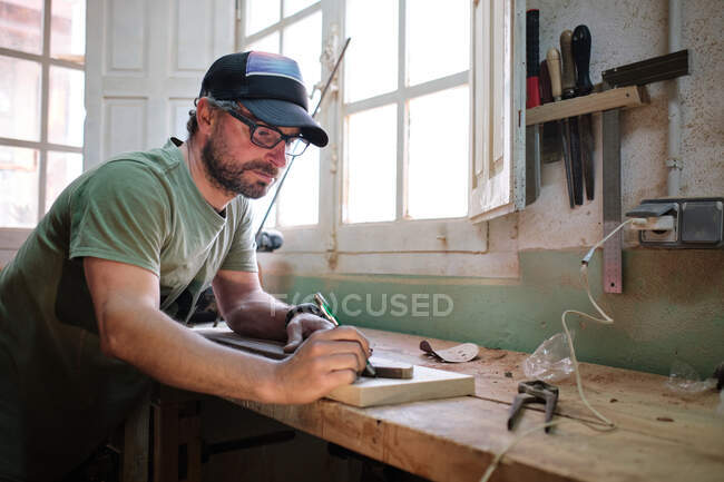 Side view of bearded workman in eyeglasses and cap tracing skateboard form with marker on wooden plank in workshop — Stock Photo