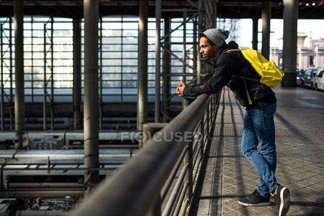 Seitenansicht eines männlichen Reisenden mit Rucksack, der am Geländer eines Durchgangs über Zügen am Bahnhof steht — Stockfoto