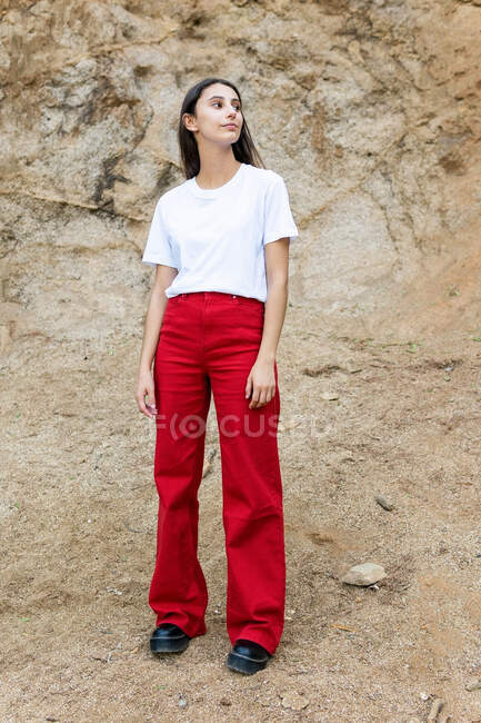 Young contemplative female adolescent in white t shirt and red jeans looking away while standing on rough land against mount — Stock Photo
