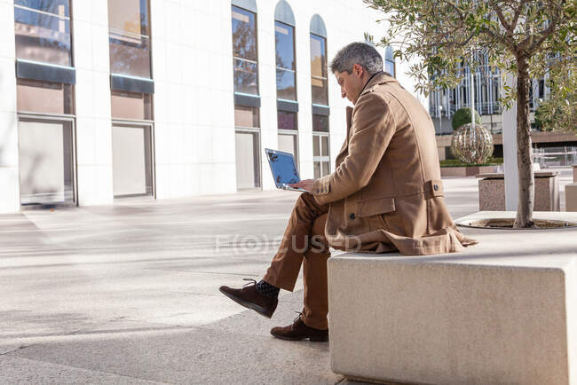 Full body of concentrated young male in formal wear sitting on bench and browsing laptop while working on project in city — Stock Photo