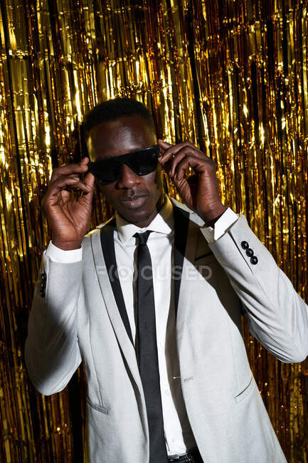 Cool African American male in jacket and tie holding on to his sunglasses against tinsel while celebrating New Years Eve — Stock Photo