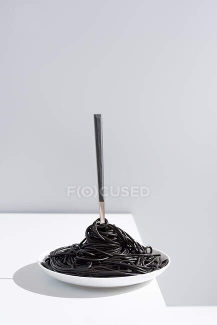 Fourchette en acier inoxydable dans un bol plein de spaghettis noirs à l'encre de seiche sur table blanche en studio sur fond gris — Photo de stock