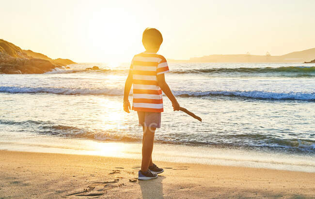 Back view full length of unrecognizable boy with stick standing on wet sandy shore washed by waving blue sea at sundown — Stock Photo