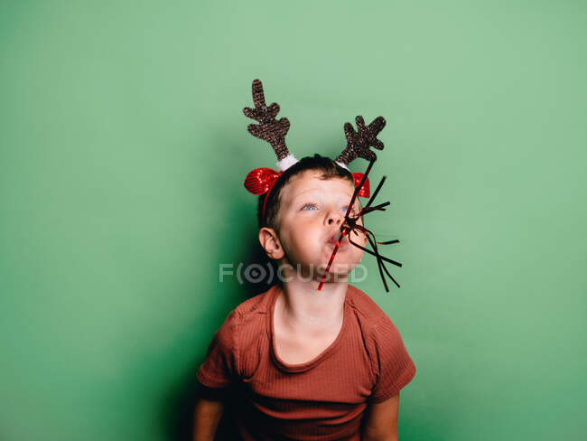 Niño usando cuernos de reno diadema y fiesta festiva soplador en la boca de pie sobre fondo verde y mirando hacia otro lado - foto de stock