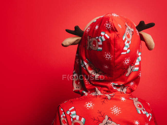 Vue arrière du petit enfant mignon méconnaissable en pyjama de Noël à capuchon avec des cerfs debout sur fond rouge — Photo de stock