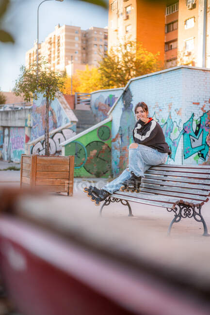 Corpo inteiro de jovem mulher positiva vestindo capuz preto casual e jeans azul claro com fendas e lâminas de rolo sentado na parte de trás do banco de madeira no parque de skate — Fotografia de Stock