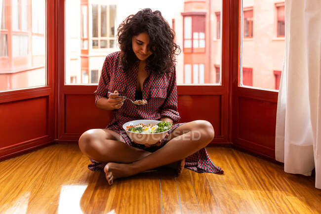Молодая женщина с вьющимися волосами сидит со скрещенными ногами и ест супер миску еды с куриным горошком нарезанные яйца брокколи — стоковое фото