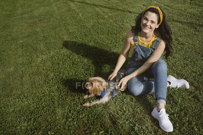 Hochwinkelvoller Körper einer glücklichen jungen Frau, die mit überkreuzten Beinen auf dem Rasen sitzt und den Yorkshire Terrier streichelt, während sie in die Kamera schaut — Stockfoto