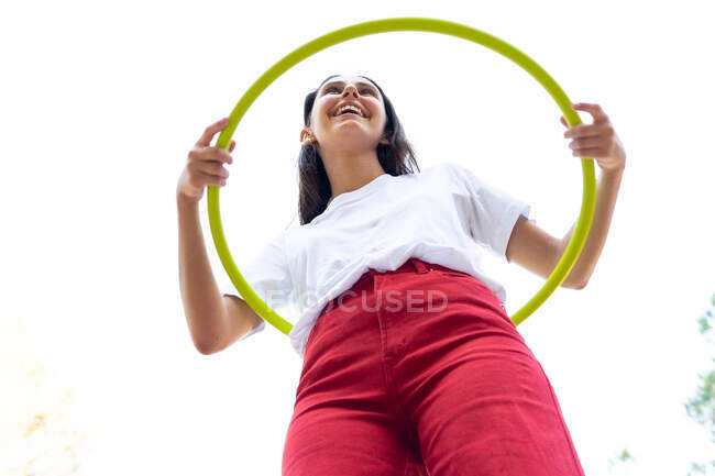 From below of cheerful female teen in red jeans twirling hula hoop while having free time in park — Stock Photo