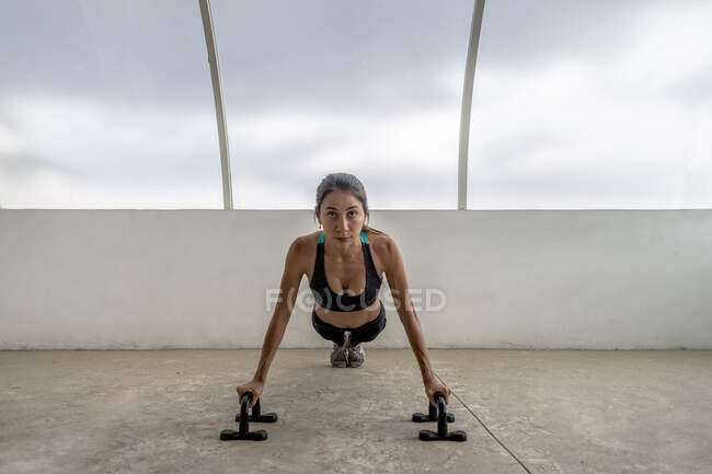 Fit focused ethnic sportswoman in active wear exercising on push up bars while looking forward — Stock Photo