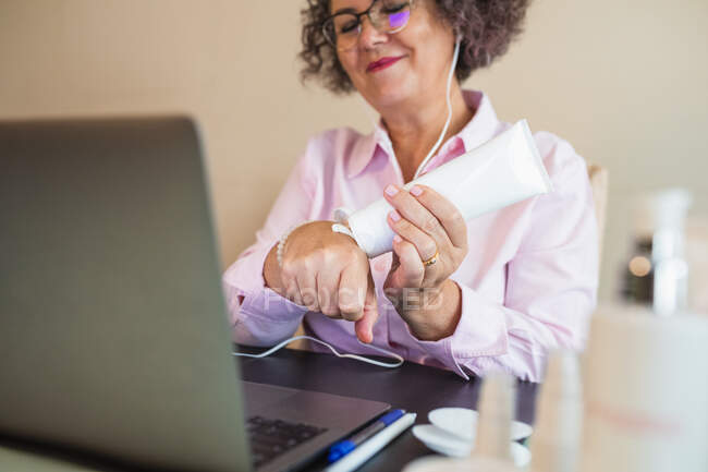 Crop content elderly female entrepreneur in earphones applying moisturizing cream on hand while having video call on laptop in office — Stock Photo