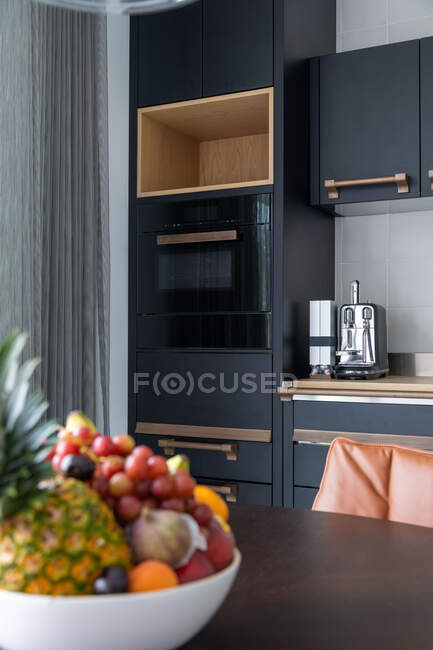 Чаша со свежими фруктами на деревянном столе на современной кухне с минималистской черной мебелью при дневном свете — стоковое фото
