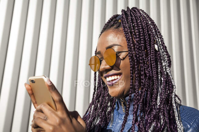 Joven mujer afroamericana alegre y elegante con trenzas afro con chaqueta de moda y gafas de sol navegando por las redes sociales en el teléfono móvil mientras está de pie cerca de la pared del edificio urbano - foto de stock