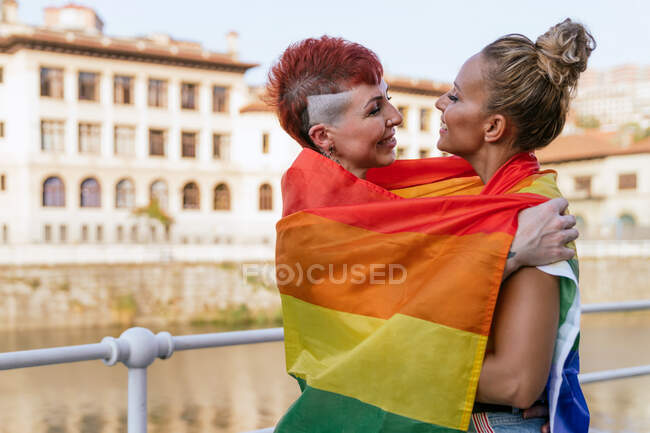 Крутая татуированная женщина с ирокезом и флагом ЛГБТК обнимает подругу с закрытыми глазами против канала в городе — стоковое фото