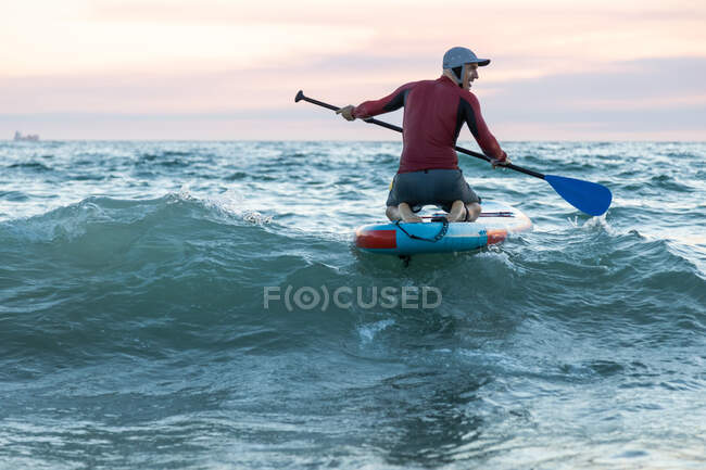 Задний вид на неузнаваемого серфера в гидрокостюме и шляпе на гребной доске для серфинга на берегу моря — стоковое фото